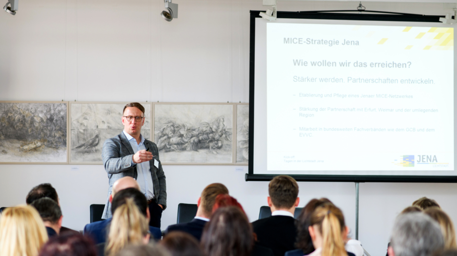 Quelle: JenaKultur, Foto: Christoph Worsch Redner präsentiert die Tagesagenda auf einer Leinwand den Teilnehmern